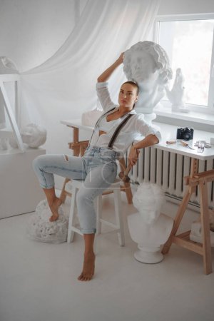 Photo for Talented female sculptor dressed in a white unbuttoned shirt and jeans with suspenders, holding a hammer and posing on a chair in her sculptor workshop surrounded by sculptures - Royalty Free Image
