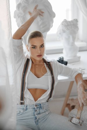 Photo for Stunning woman in a disheveled white shirt and jeans with suspenders, confidently seated on a chair in her workshop, holding a hammer and surrounded by her intricate sculptures - Royalty Free Image