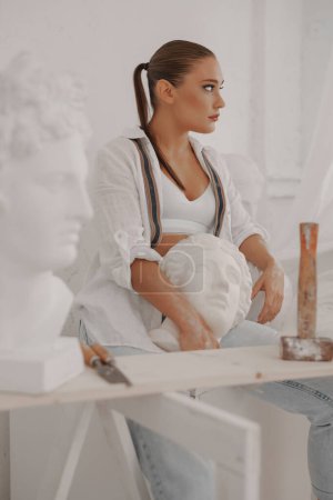 Téléchargez les photos : Sculptrice talentueuse dans une chemise blanche et un jean, tenant un buste grec antique dans son atelier, entourée d'outils. Un mélange parfait d'artisanat et d'art - en image libre de droit