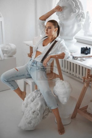 Téléchargez les photos : Une jolie sculptrice vêtue d'une chemise blanche lâche et d'un jean avec des bretelles, assise sur un tabouret dans son atelier de sculpteurs - en image libre de droit