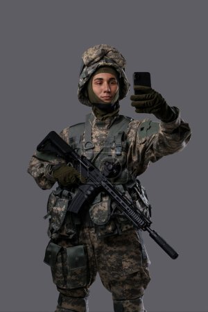 Foto de Militar con un arma y un teléfono móvil toma una selfie o tiene un video chat, mostrando un lado casual y moderno de la vida de un soldado - Imagen libre de derechos