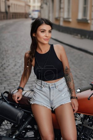Téléchargez les photos : Superbe brune assise sur sa moto rétro contre au milieu d'une rue en Europe - en image libre de droit