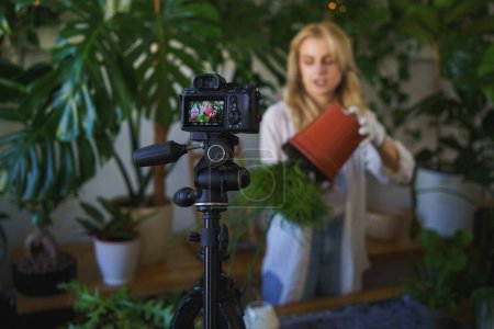 Téléchargez les photos : Blogueuse passionnée de plantes en chemise blanche et en jean, souriante à la lumière naturelle de son appartement urbain rempli de plantes - en image libre de droit