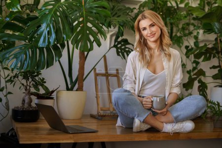 Téléchargez les photos : Belle femme blonde profite d'une tasse de thé tout en étant assis à côté de son ordinateur portable sur une table entourée de plantes en pot - en image libre de droit