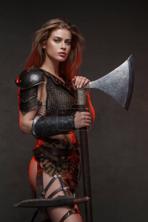 Impresionante mujer vikinga vestida con una parte superior de malla de malla y falda de piel posa con un hacha de dos manos iluminada por la iluminación del borde rojo 