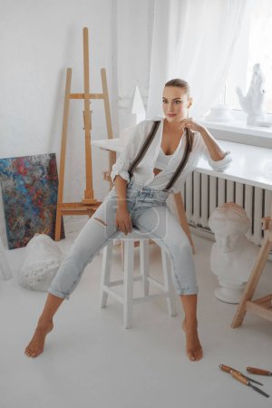 Téléchargez les photos : Une jolie sculptrice vêtue d'une chemise blanche lâche et d'un jean avec des bretelles, assise sur un tabouret dans son atelier de sculpteurs - en image libre de droit