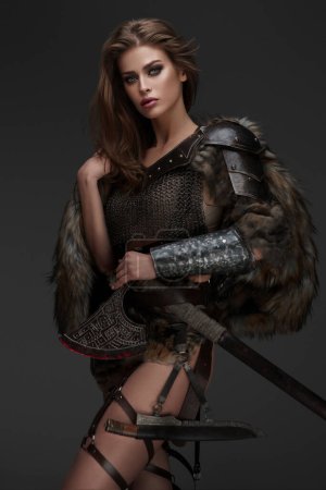 Téléchargez les photos : Un joli modèle à thème viking en armure de maille et fourrure, tenant une hache sur fond gris - en image libre de droit