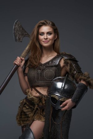 Photo for Smiling beautiful model Viking woman wearing a chainmail top and fur posing with an axe and helmet against a gray background - Royalty Free Image