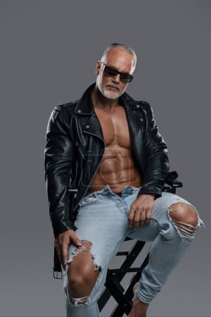 Schöne durchtrainierte männliche Modell mit einem stilvollen grauen Bart mit Sonnenbrille, trägt eine schwarze Lederjacke und zerrissene Jeans, enthüllt seinen muskulösen Körper und nackten Oberkörper, während er auf einem Studiostuhl posiert