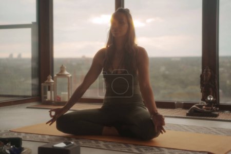 Foto de Mujer con atuendo deportivo verde practicando yoga y meditación en pose de loto sobre un paisaje urbano panorámico en un apartamento - Imagen libre de derechos