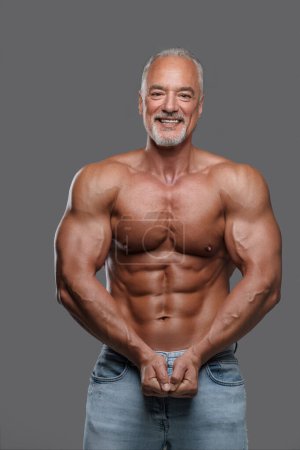 Foto de Hombre mayor atractivo con un físico muscular, elegante barba gris y vaqueros rasgados, sonriendo y posando sin camisa sobre un fondo gris - Imagen libre de derechos