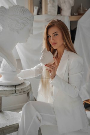 Photo for Exquisite young woman, with a models appearance, wearing a stylish white suit and seductive bra top, holding piece of plaster mask while sitting in a sculptors studio near ancient Greek bust - Royalty Free Image