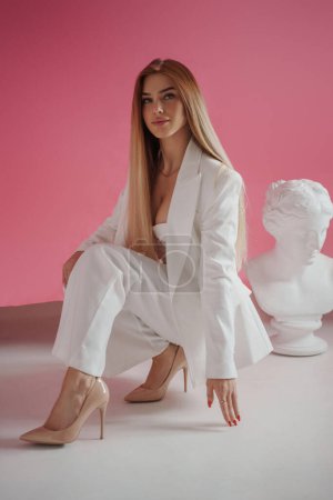 Elegant model wearing a white suit and sexy bra, posing next to an ancient Greek bust in a glamorous photoshoot in a pink studio