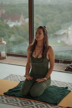 Téléchargez les photos : Femme en tenue de sport verte pratiquant le yoga et la méditation contre les fenêtres panoramiques dans un appartement - en image libre de droit
