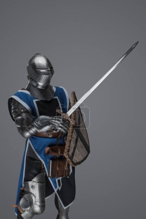 Foto de Brave medieval warrior dressed in armor and blue surcoat waving his sword energetically in battle, against a gray backdrop - Imagen libre de derechos