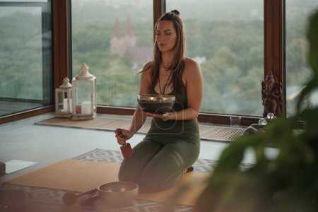 Téléchargez les photos : Femme en tenue de sport pratiquant des activités spirituelles dans un environnement paisible, méditant avec un bol tibétain chantant dans un appartement donnant sur un horizon panoramique de la ville - en image libre de droit
