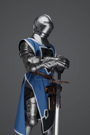 A medieval soldier garbed in blue surcoat and armor holding a sword with a still, statue-like pose, against a gray background