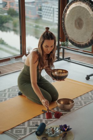 Téléchargez les photos : Une atmosphère sereine en tant que femme se livre à des pratiques spirituelles, méditant avec un bol tibétain chantant dans un appartement de la ville dans un contexte urbain pittoresque - en image libre de droit