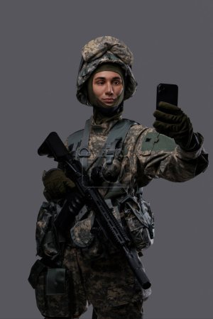 Téléchargez les photos : Jeune homme en uniforme militaire et casque tenant un fusil et un smartphone, posant pour un selfie ou ayant un appel vidéo sur un fond gris - en image libre de droit