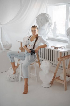 Foto de Hermosa escultora vestida con una camisa blanca desabotonada y jeans con tirantes, sosteniendo una cámara mientras está sentada en una silla en su taller de escultor - Imagen libre de derechos