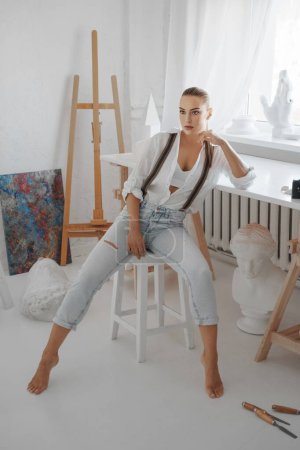 Photo for Beautiful sculptor, wearing a white unbuttoned shirt and jeans with suspenders, sits on a chair in her workshop - Royalty Free Image
