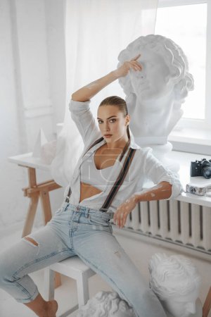 Photo for An attractive female sculptor dressed in a loose white shirt and jeans with suspenders, while sitting on a stool in her sculptors studio - Royalty Free Image