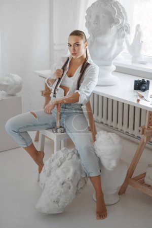 Foto de Talentosa escultora vestida con una camisa blanca desabotonada y jeans con tirantes, sosteniendo un martillo y posando en una silla en su taller de escultores rodeada de esculturas - Imagen libre de derechos