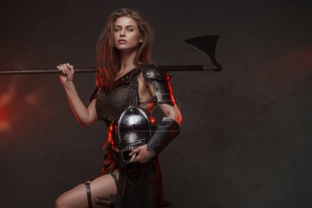 Impresionante mujer vikinga vestida con una parte superior de malla de malla y piel con un hacha de dos manos iluminada por la iluminación del borde rojo