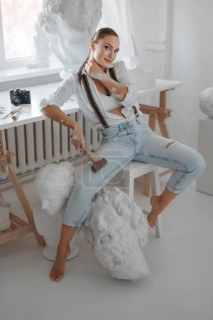 Foto de Talentosa escultora vestida con una camisa blanca desabotonada y jeans con tirantes, sosteniendo un martillo y posando en una silla en su taller de escultores rodeada de esculturas - Imagen libre de derechos