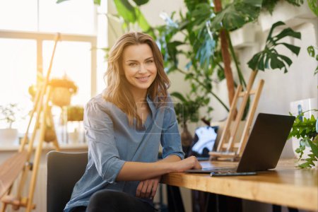 Téléchargez les photos : Belle femme indépendante souriante en chemise bleue travaillant sur ordinateur portable dans une pièce ensoleillée remplie de plantes - en image libre de droit
