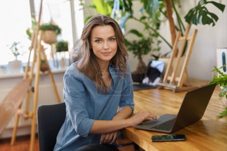 Photo for A charming smiling freelancer woman in a blue shirt working on a laptop in a plant-filled room - Royalty Free Image