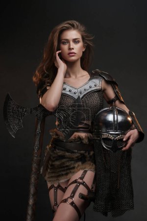 Schöne Wikinger Krieger Modell posiert mit einer leistungsstarken Axt und Helm, die Stärke und Weiblichkeit in einem mittelalterlich inspirierten Kostüm vor einem strukturierten Hintergrund zu präsentieren