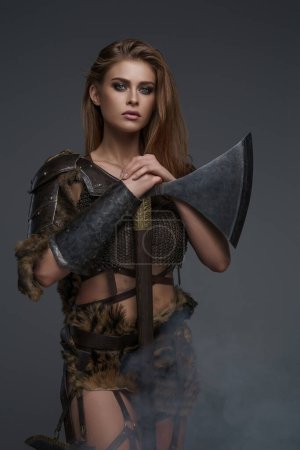 Impresionante modelo vikingo vestido con armadura de malla y piel, posando con un hacha contra una pared gris
