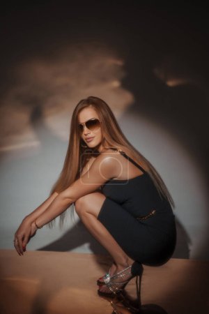 Foto de Impresionante chica en vestido de noche negro y soleados, golpeando una pose sentada en posición de sentadilla en un piso de oro reflectante, con una magnífica iluminación creando un telón de fondo fascinante - Imagen libre de derechos