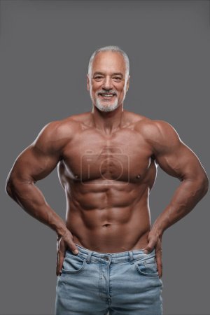 Photo for Attractive older man with a muscular physique, stylish gray beard and ripped jeans, smiling and posing shirtless against a gray backdrop - Royalty Free Image