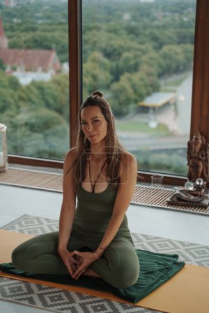 Photo for An athletic woman in green workout clothes doing yoga in lotus position with a backdrop of panoramic windows in an apartment - Royalty Free Image