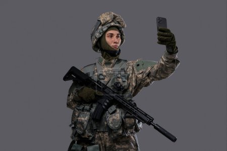 Foto de Soldado en la celebración de un arma y un teléfono móvil, que parece tomar una selfie o hacer una videollamada en un fondo gris - Imagen libre de derechos
