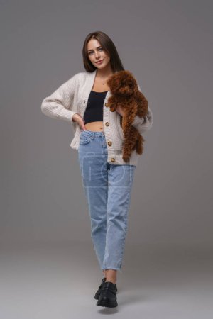 Photo for Full-length portrait of a captivating brunette in casual wear holding her brown toy poodle against a gray studio background - Royalty Free Image