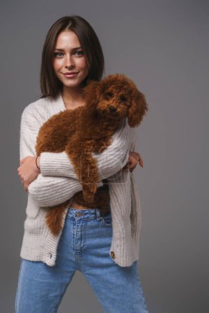 Téléchargez les photos : Charmante fille dans un pull blanc tenant son caniche jouet brun bien-aimé sur un fond de studio gris - en image libre de droit