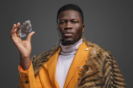Photo for Dapper dark-skinned man adorned in a stunning yellow blazer and draped in a wild animal pelt, showcasing an enormous diamond on a gray background - Royalty Free Image