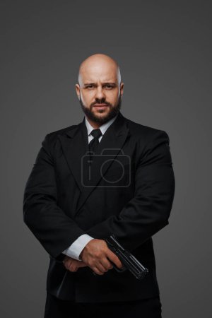 Téléchargez les photos : Un agent de sécurité chauve en costume tient une arme, prêt à protéger, dans un décor de studio gris - en image libre de droit
