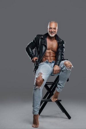 Photo for Masculine and stylish male model with striking physique, grey beard, and bare chest, wearing black leather jacket and ripped jeans, sitting on a studio chair against a neutral grey backdrop - Royalty Free Image