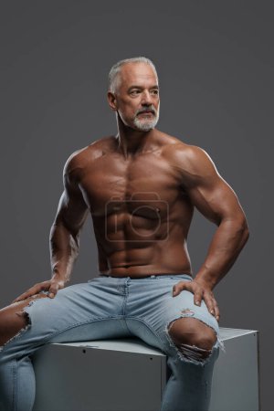 Foto de Modelo masculino musculoso encantador de edad madura, con una elegante barba gris y un torso desnudo que lleva vaqueros rasgados, posando en un cubo gris en un estudio fotográfico sobre un fondo gris - Imagen libre de derechos