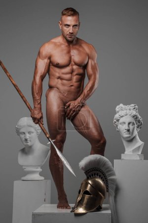 Ein muskulöses und attraktives männliches Modell posiert mit einem Speer, der seine Geschlechtsteile bedeckt, und steht auf einem Sockel mit einem griechischen Helm, umgeben von antiken griechischen Skulpturen
