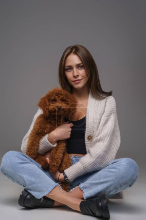 Attractive young brunette in white top and denim shorts, seated on the floor, cradling her brown toy poodle, against a gray studio backdrop