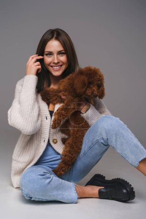 Photo for Portrait of a smiling brunette sitting on the floor in casual attire, embracing her cherished toy poodle in a gray studio setting - Royalty Free Image