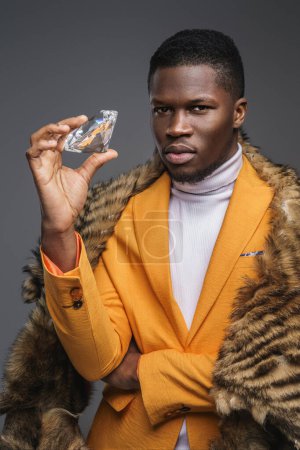 Photo for A stylish ebony gentleman sporting a yellow jacket and fur shawl, flaunting a massive diamond against a neutral gray backdrop - Royalty Free Image