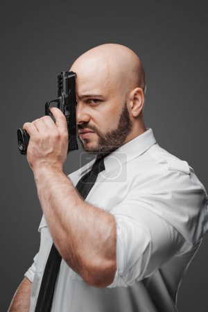 Photo for Dramatic portrait of a bald, bearded man in white shirt and tie, pressing a pistol to his face, symbolizing detective, bodyguard, or security themes against a gray backdrop - Royalty Free Image