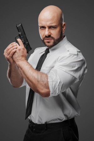 Photo for Bald, bearded man in white shirt and tie holds a pistol, epitomizing a detective, bodyguard, or security professional against a gray studio background - Royalty Free Image