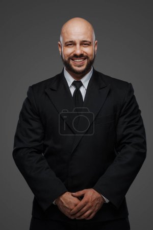 Téléchargez les photos : Heureux gentleman barbu dans un élégant ensemble noir pose avec un sourire sur un fond neutre - en image libre de droit
